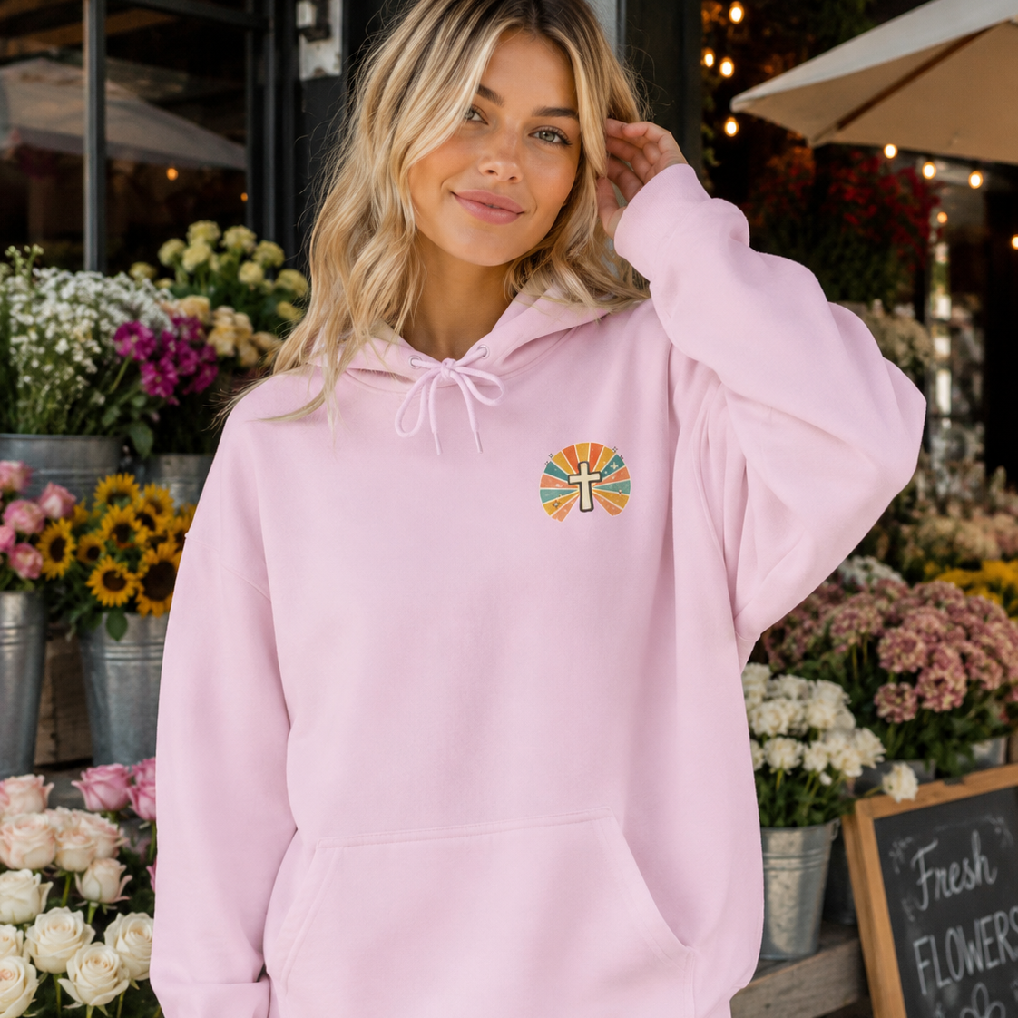 A woman wearing a light pink pullover hoodie with a small colorful circular cross graphic on the left chest, standing in a flower shop.