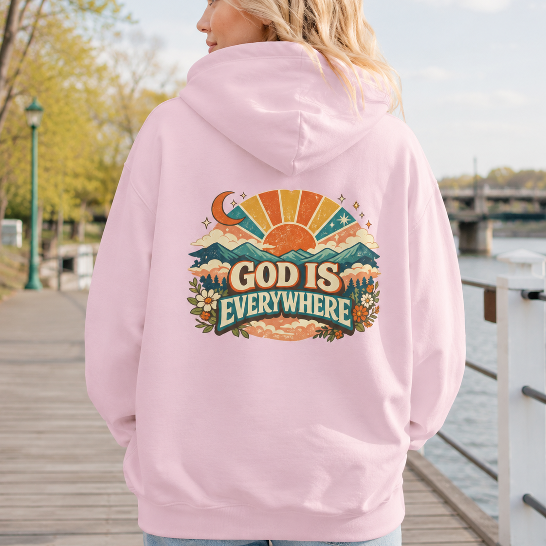A woman viewed from behind wearing a light pink hoodie with a large colorful graphic on the back that reads “GOD IS EVERYWHERE,” standing on a wooden boardwalk outdoors near water and trees.