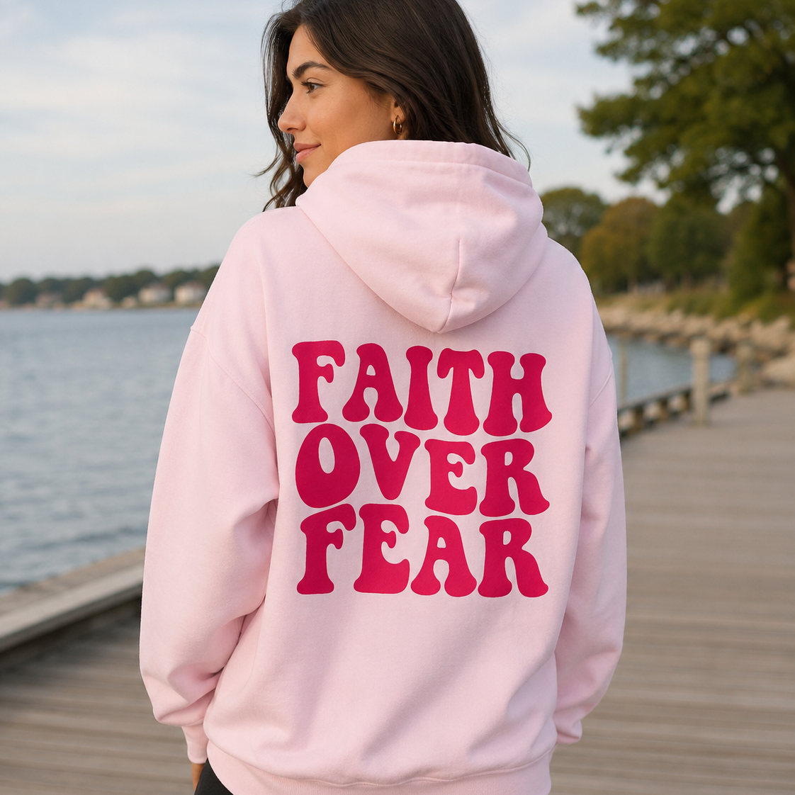 A woman wearing a light pink hoodie with the words 'FAITH OVER FEAR' printed in bold pink letters on the back, standing on a wooden boardwalk by the water.