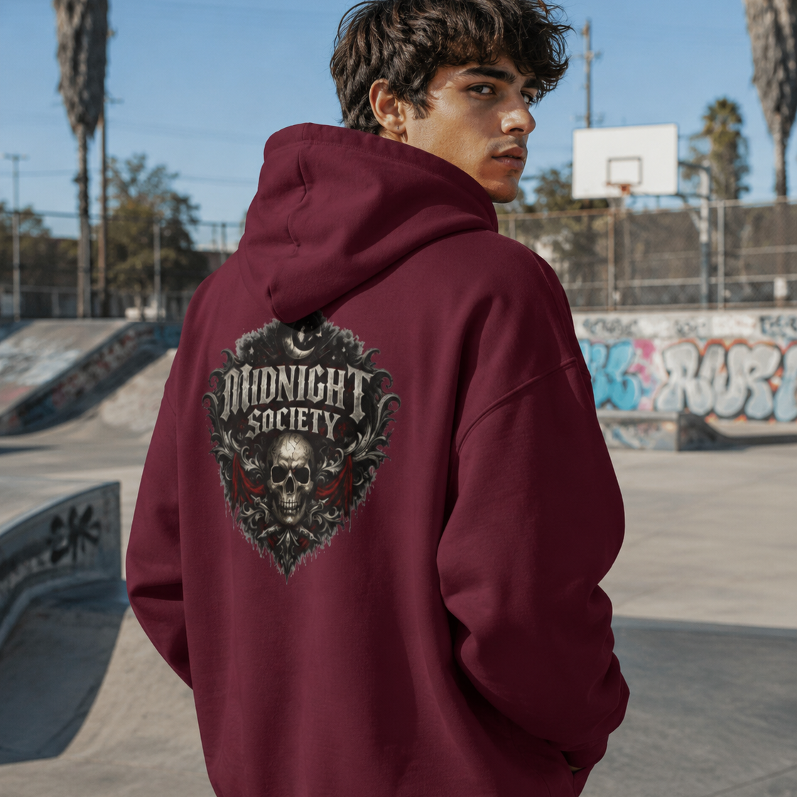 A man wearing a burgundy hoodie with a large 'Midnight Society' skull graphic on the back, standing at a concrete skatepark with palm trees and a basketball hoop in the background.
