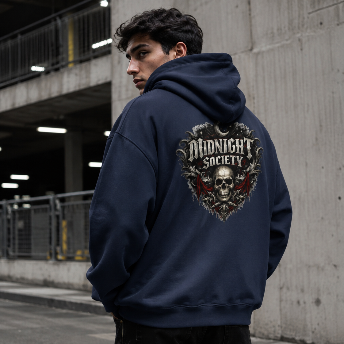 A man wearing a navy blue hoodie with a large 'Midnight Society' skull graphic on the back, standing in an urban concrete parking structure.