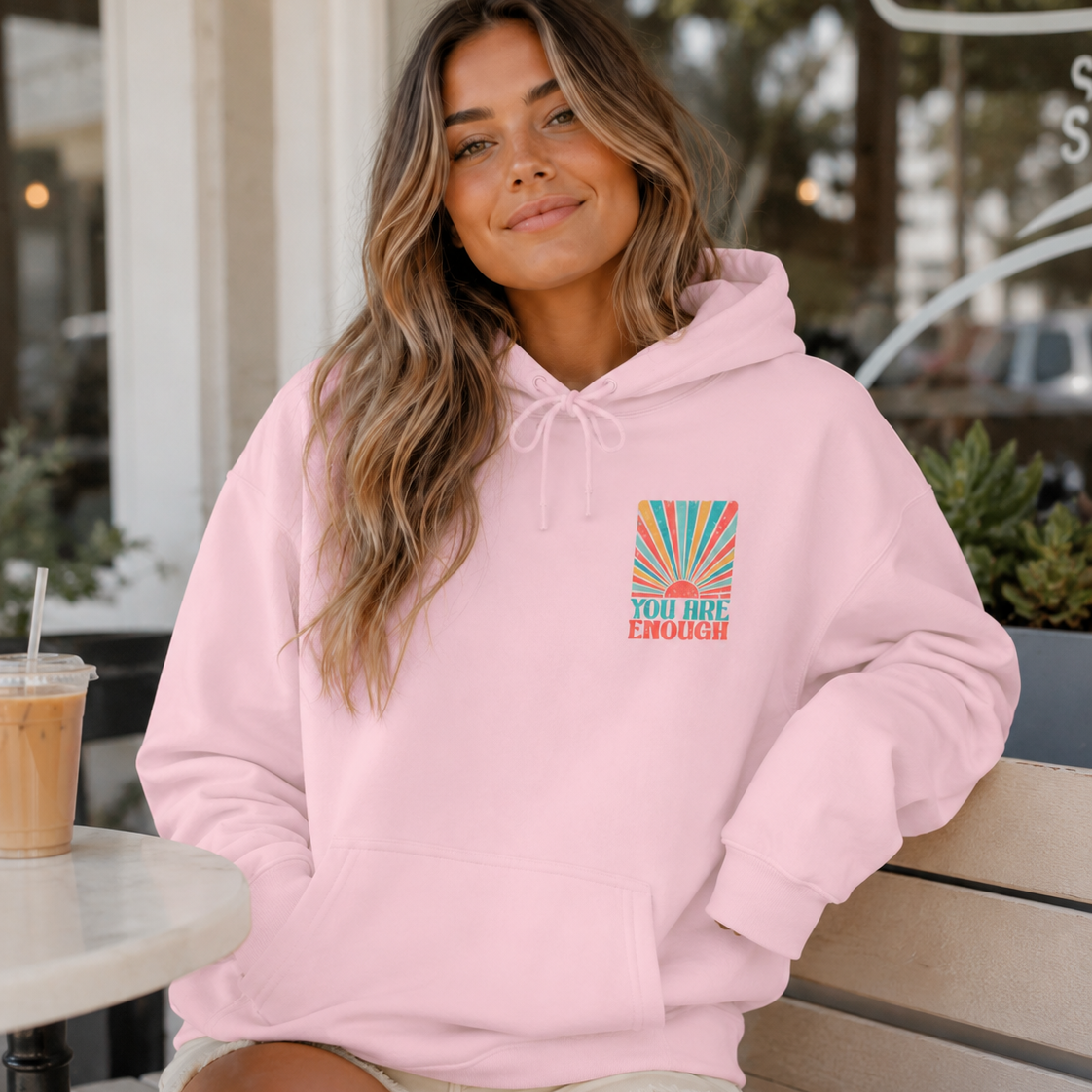 A woman sitting on a bench at a cafe wearing a light pink hoodie with a colorful graphic on the left chest that reads 'YOU ARE ENOUGH' with a sunburst design. She is wearing beige shorts and has an iced coffee on a table next to her.