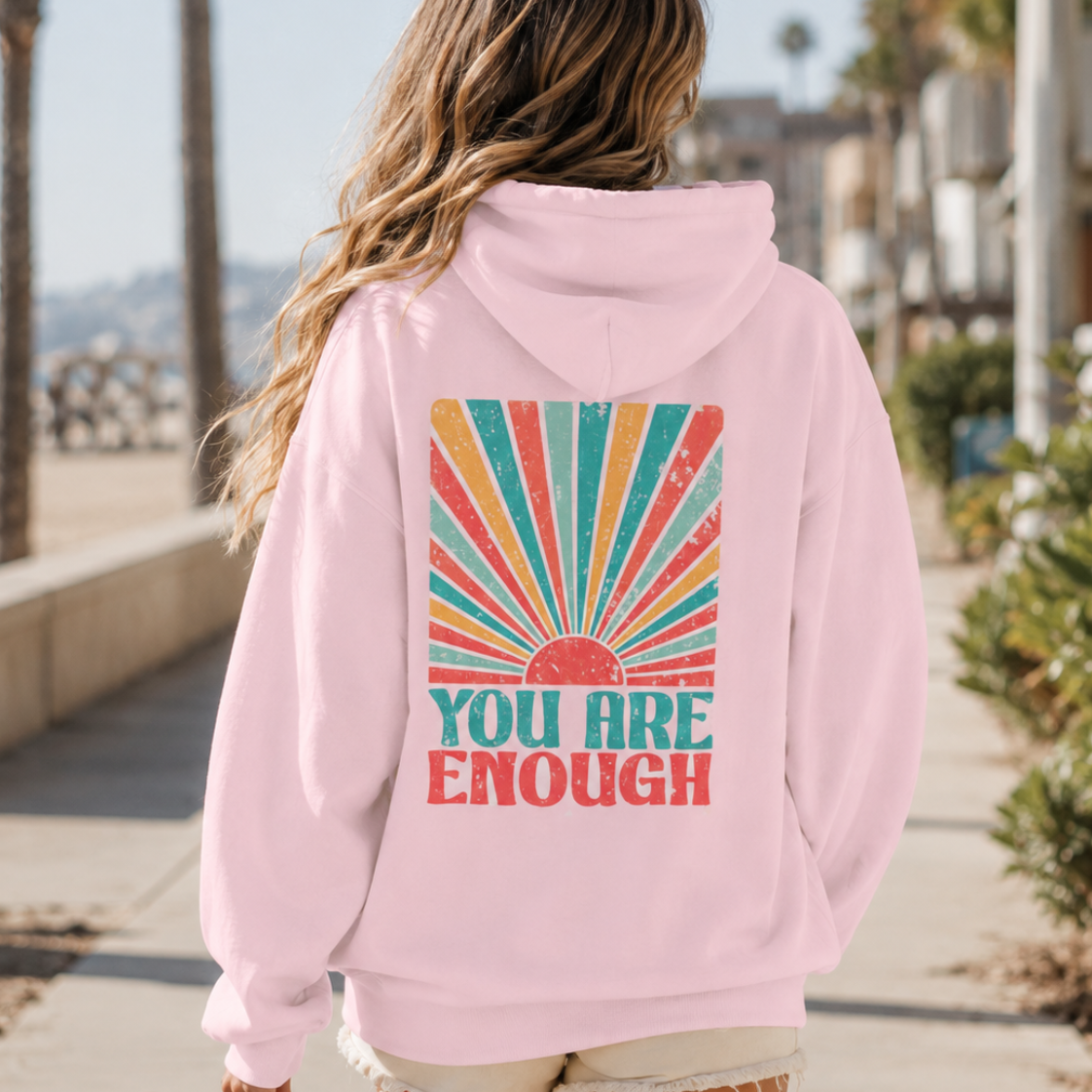 A woman viewed from behind wearing a light pink hoodie with a colorful retro sunburst graphic and the text 'YOU ARE ENOUGH' on the back, standing on a sunny beach boardwalk.