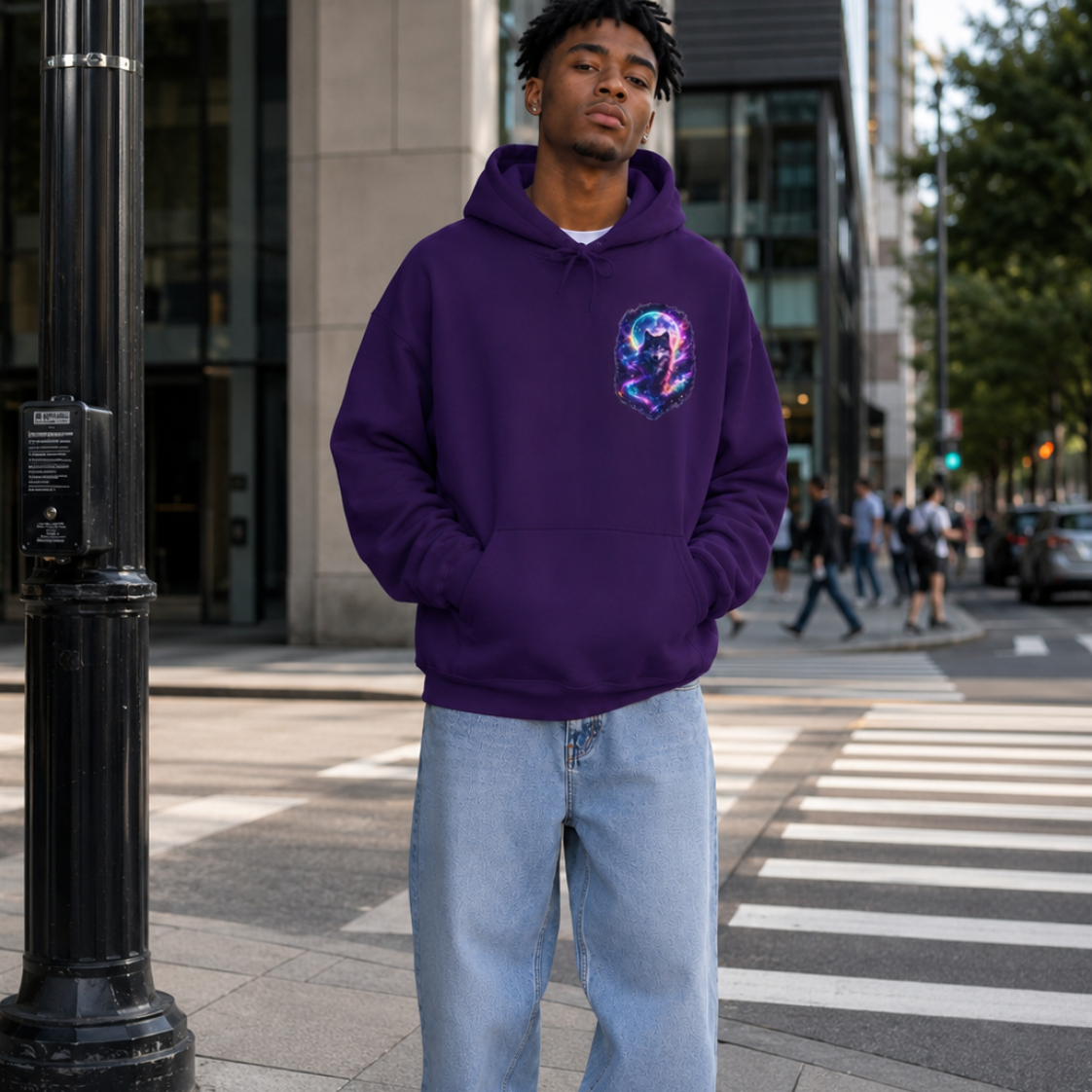 A man stands on a city sidewalk wearing a purple pullover hoodie with a colorful graphic on the left chest, light blue baggy jeans, and white sneakers.