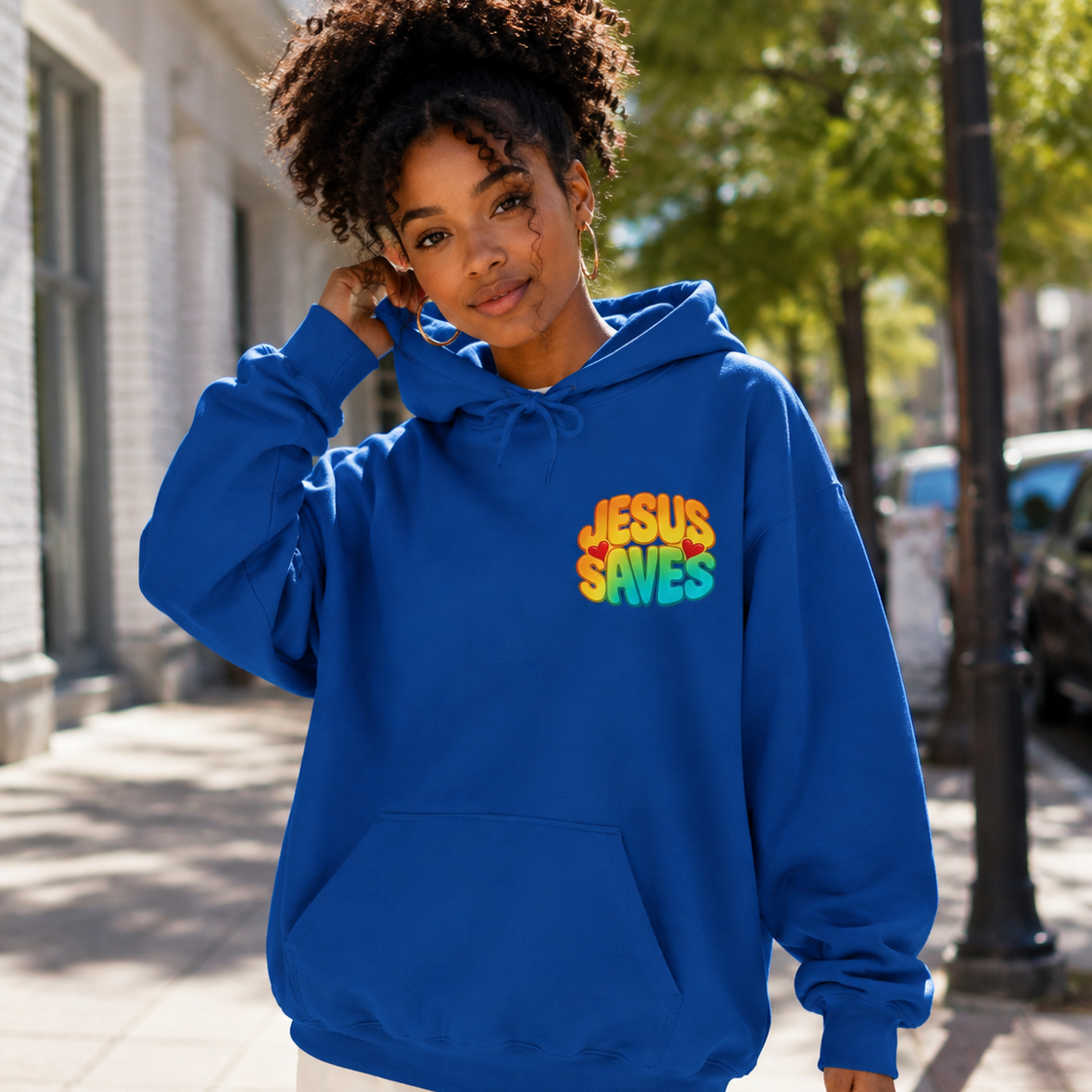 A woman wearing a bright blue pullover hoodie with the text 'JESUS SAVES' in a rainbow gradient on the chest, standing on a sunny city sidewalk.