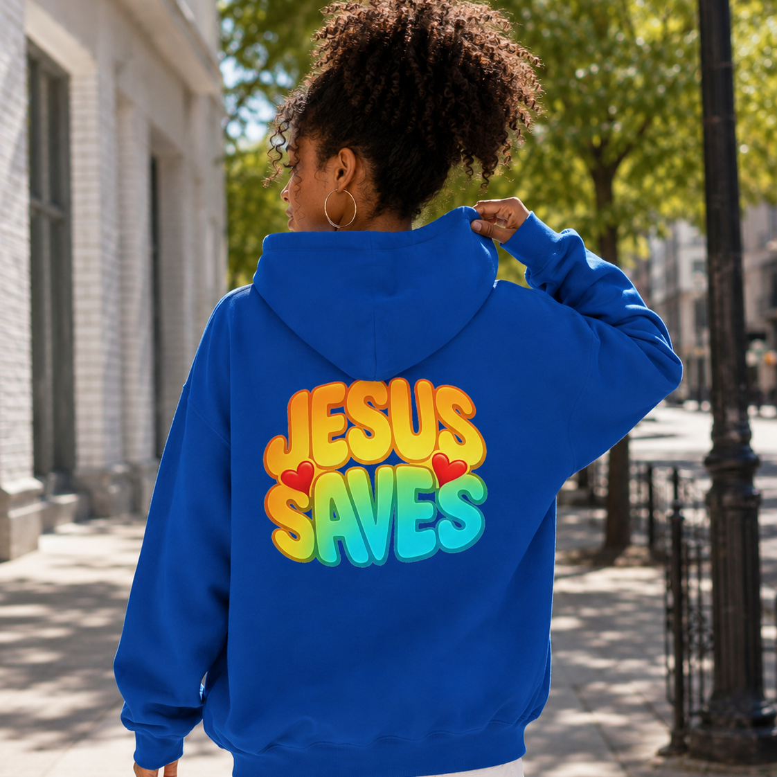 A woman viewed from behind wearing a bright blue hoodie with a colorful 'JESUS SAVES' graphic on the back, standing on a sunny city sidewalk.