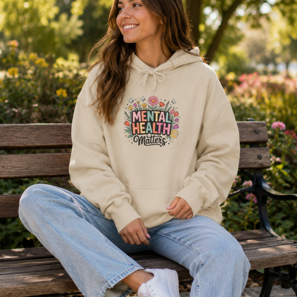 A woman sitting on a wooden park bench wearing a cream-colored hoodie with a colorful 'Mental Health Matters' graphic on the front, paired with light blue jeans and white sneakers, set against a blurred green park background.