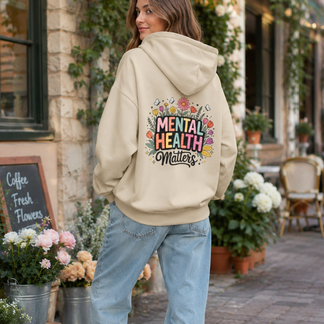 A woman stands on a cobblestone street in front of a quaint shop, wearing a beige hoodie with a colorful graphic on the back that reads 'Mental Health Matters' surrounded by flowers, paired with light blue jeans and white sneakers.