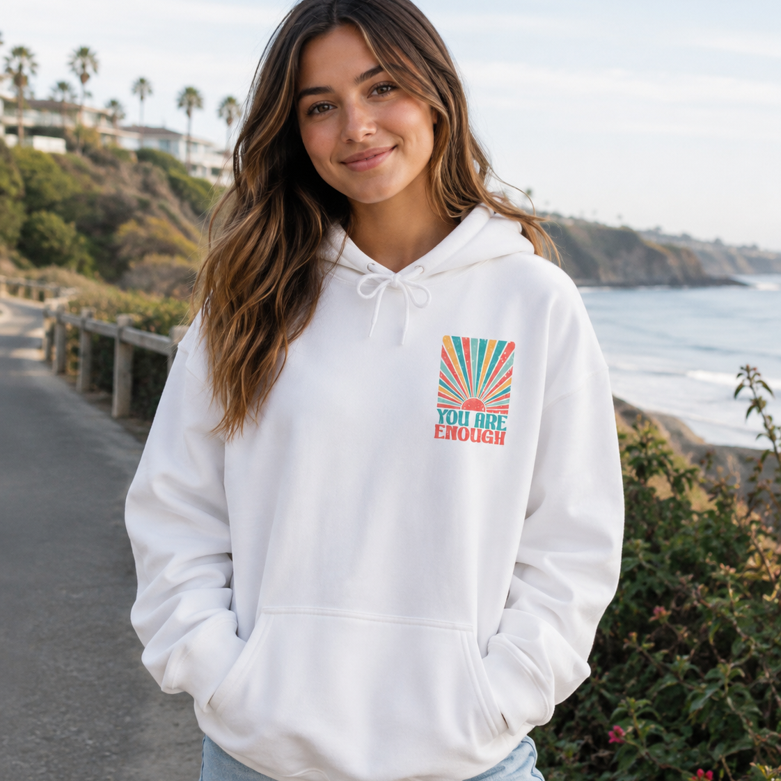 A woman wearing a white pullover hoodie with a colorful 'YOU ARE ENOUGH' graphic on the left chest, standing on a coastal path with the ocean and cliffs in the background.