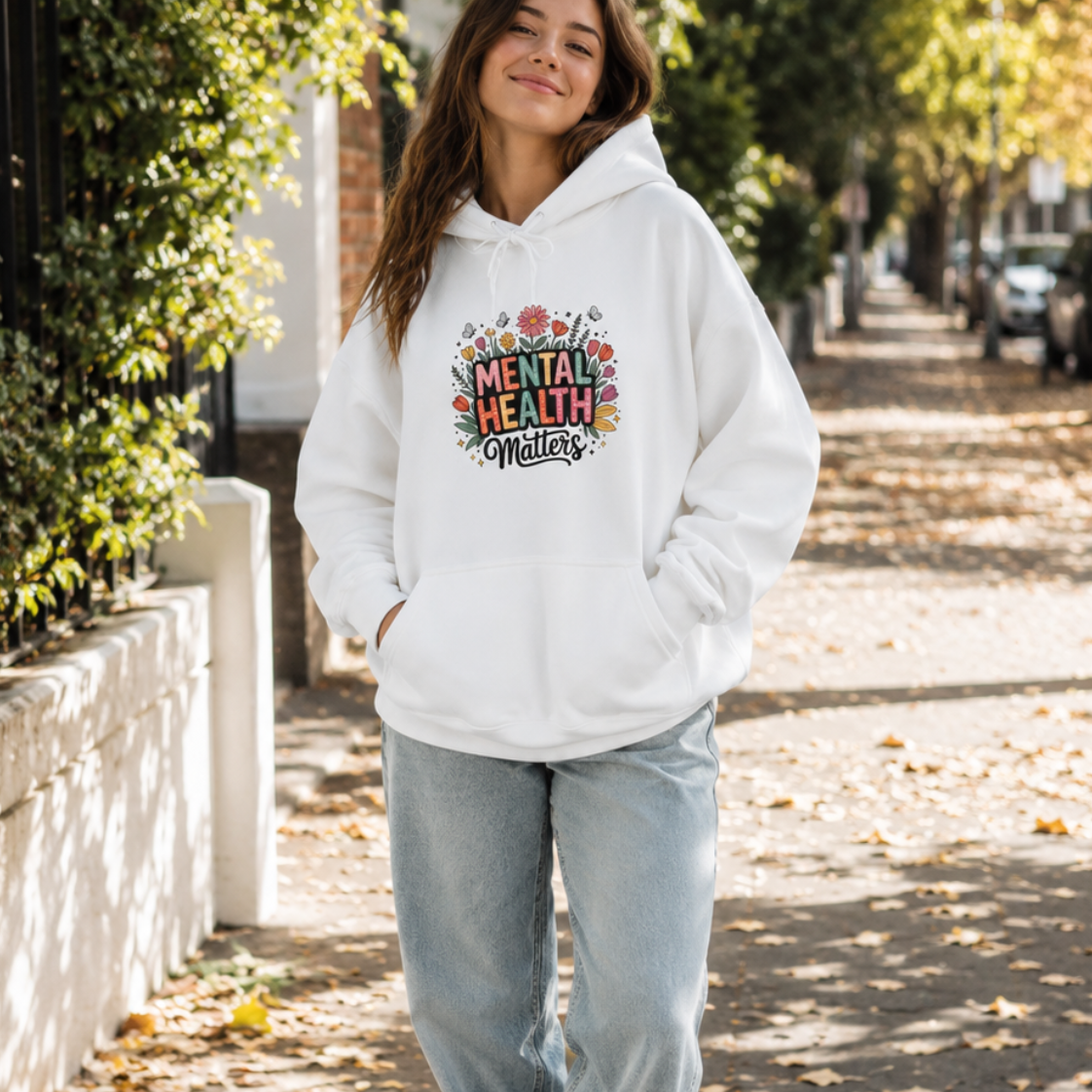A woman stands on a sunlit sidewalk wearing a white pullover hoodie with a colorful 'Mental Health Matters' graphic on the chest, paired with light blue jeans and white sneakers.