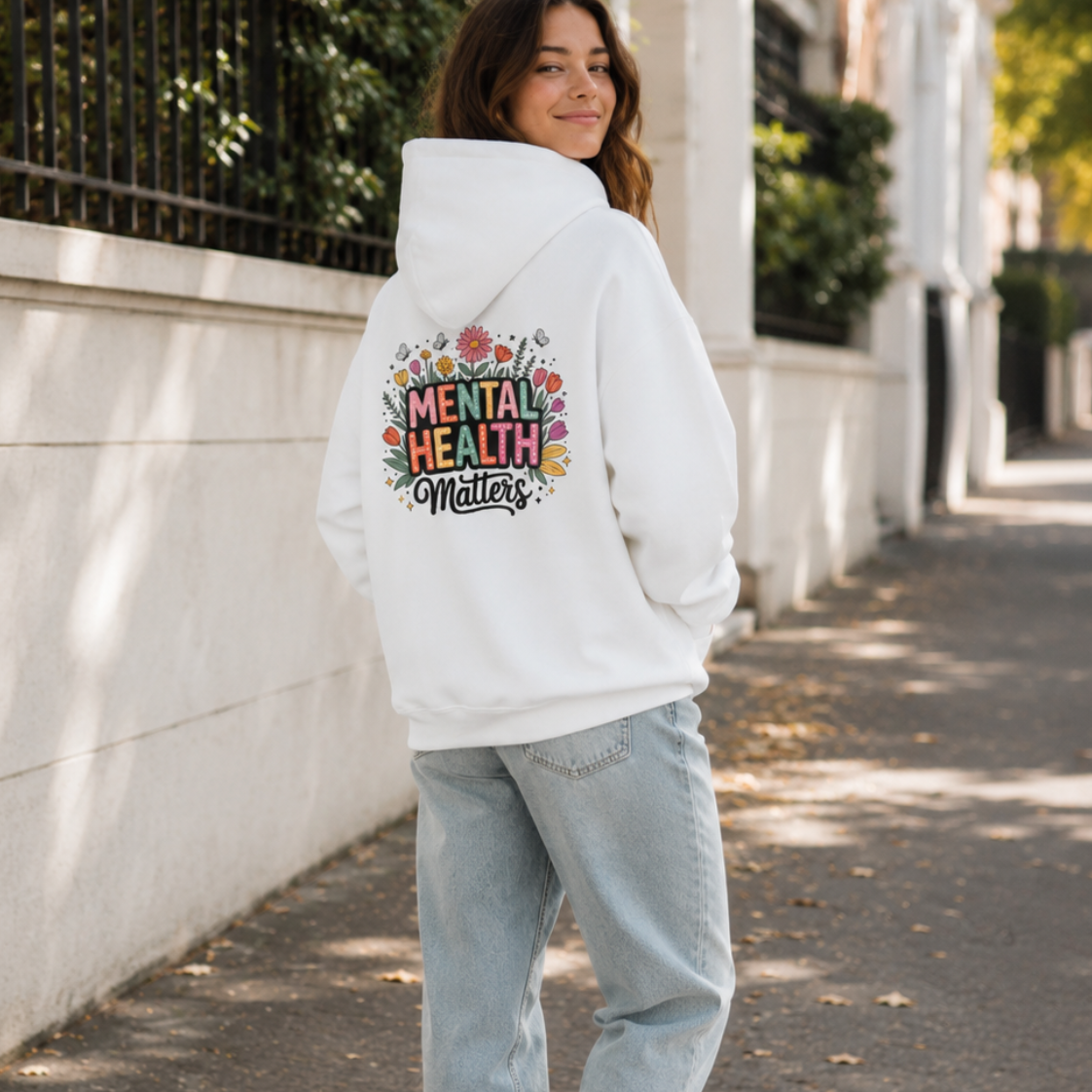 A woman stands on a sunlit sidewalk wearing a white hoodie with a colorful graphic on the back that reads 'MENTAL HEALTH MATTERS' surrounded by flowers, paired with light blue jeans and white sneakers.