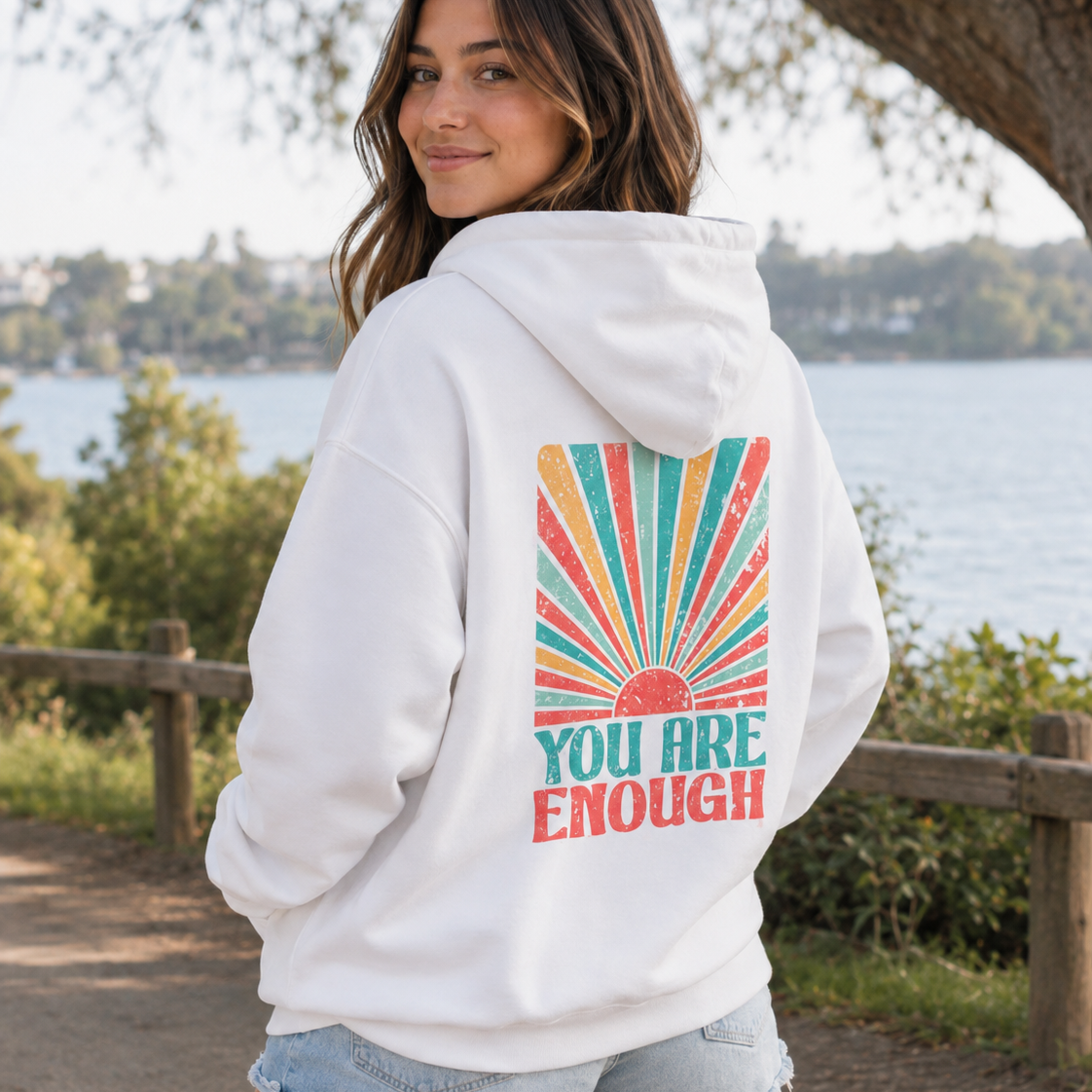 A woman wearing a white hoodie with a colorful retro sunburst graphic and the text 'YOU ARE ENOUGH' on the back, standing outdoors near a lake with trees and a wooden fence in the background.