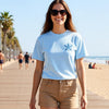 Woman walking on a wooden boardwalk by the beach wearing a light blue t-shirt with a starfish design and beige shorts.