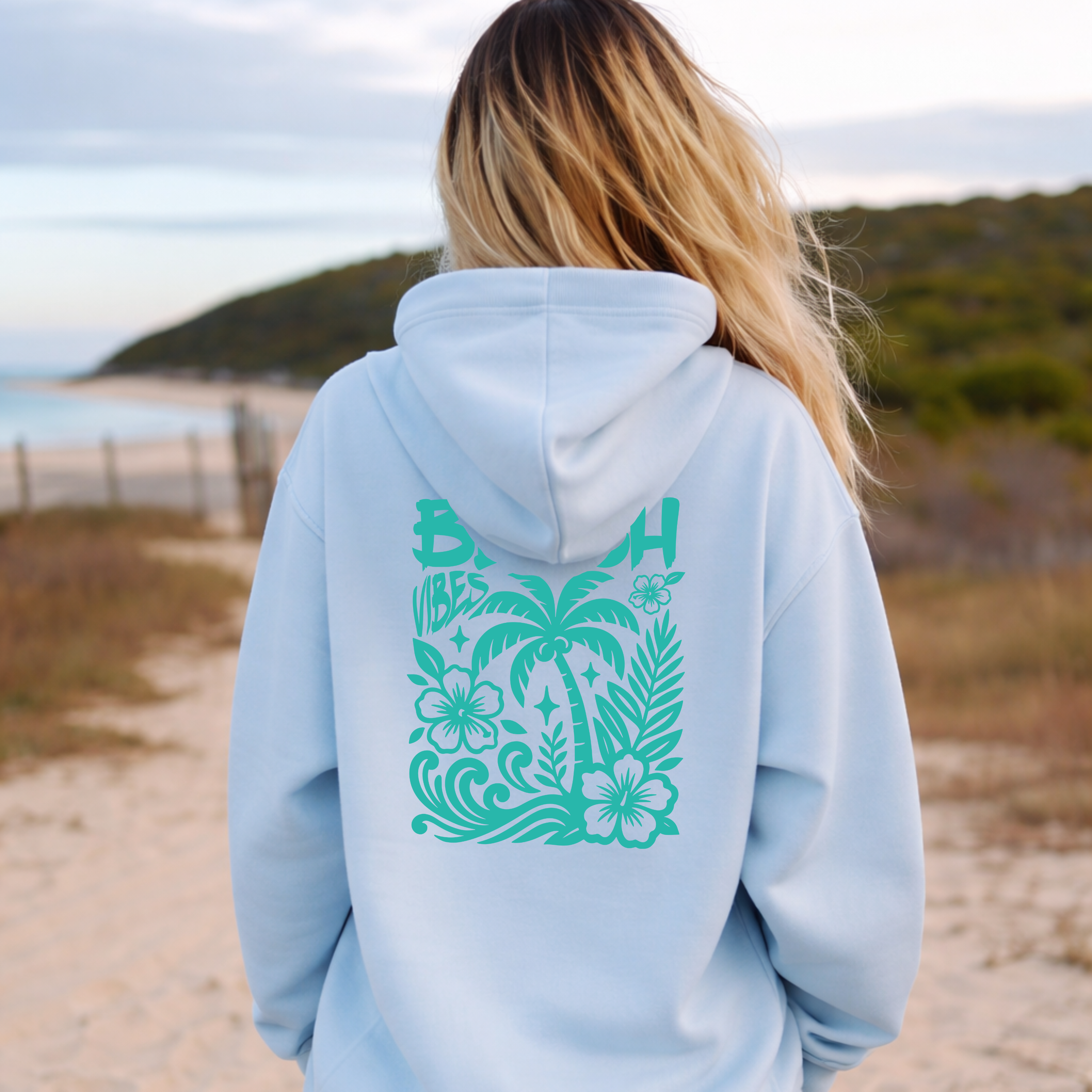Person wearing a light blue hoodie with a tropical design on the back, standing on a beach.