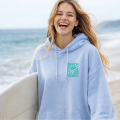Woman in a light blue hoodie with 'Beach' text and palm tree design, holding a surfboard on a beach.