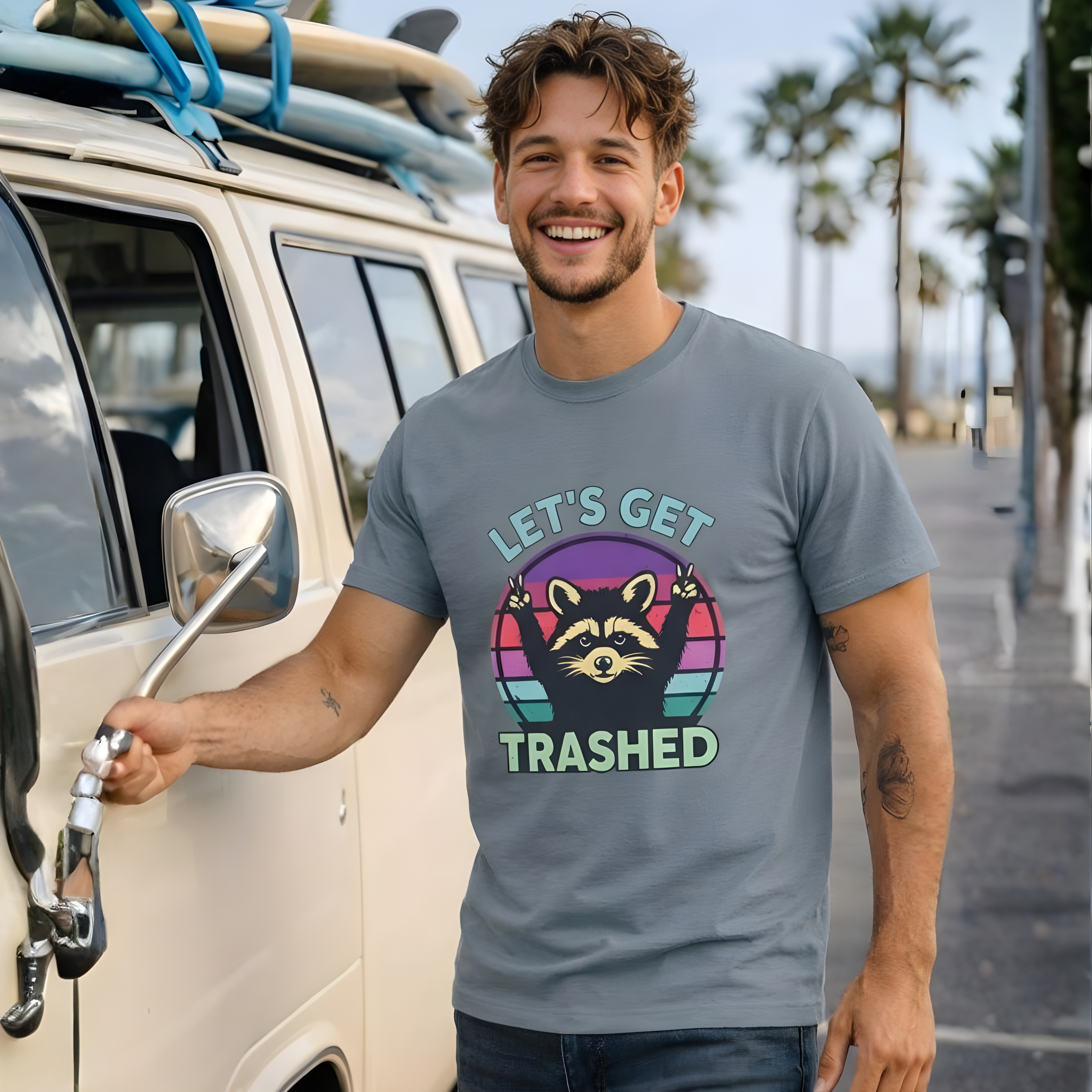 Man wearing a t-shirt with a graphic design and text, standing next to a van with surfboards on a beachside street.