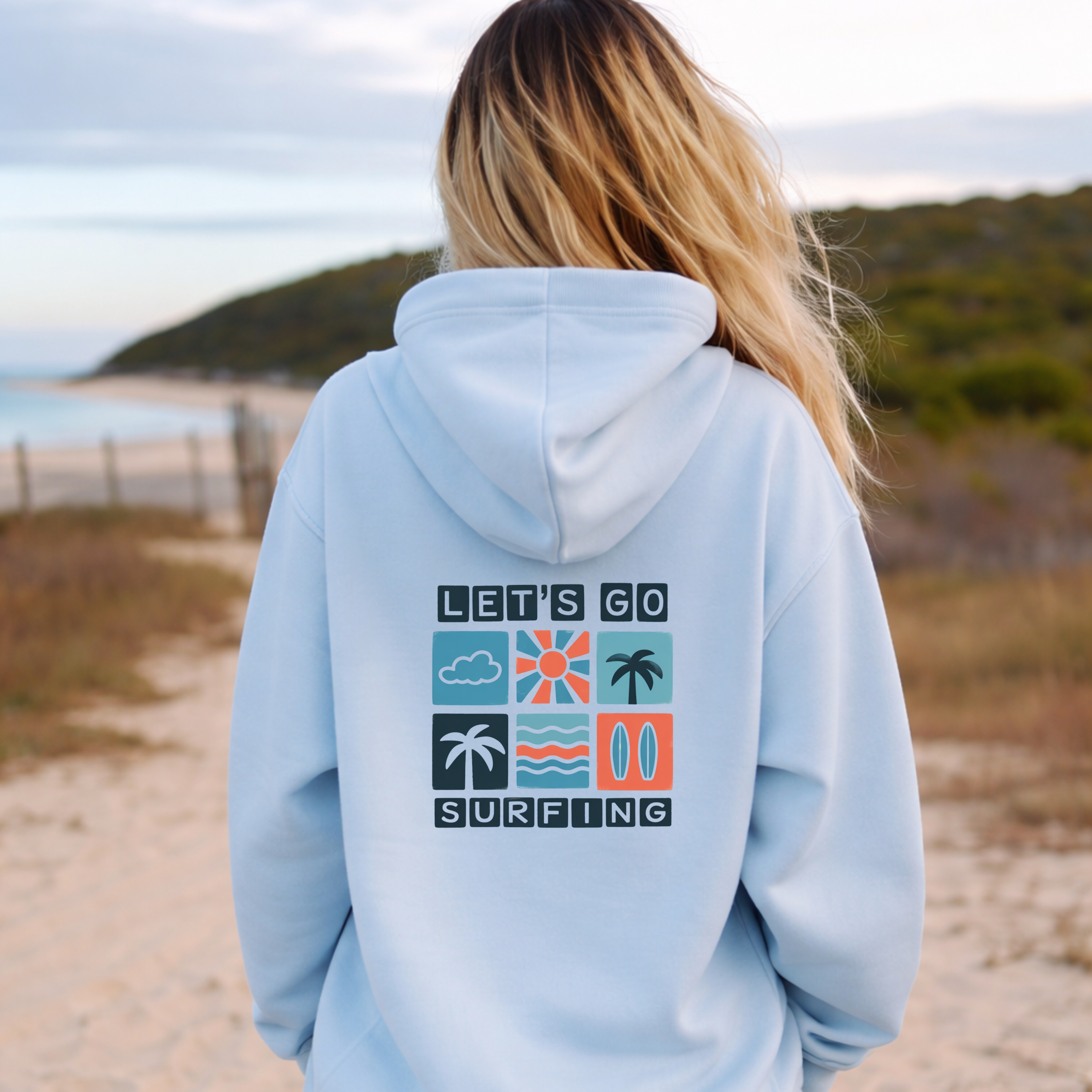 Person wearing a light blue hoodie with 'Let's Go Surfing' design on a beach.