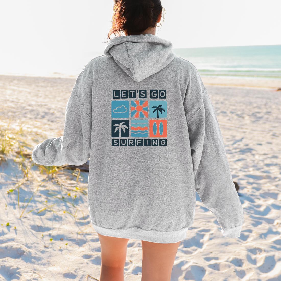 Person wearing a gray hoodie with 'Let's Go Surfing' text on a beach