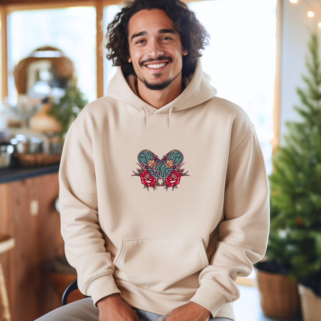Man wearing a beige hoodie with a skateboard graphic, sitting in a kitchen.
