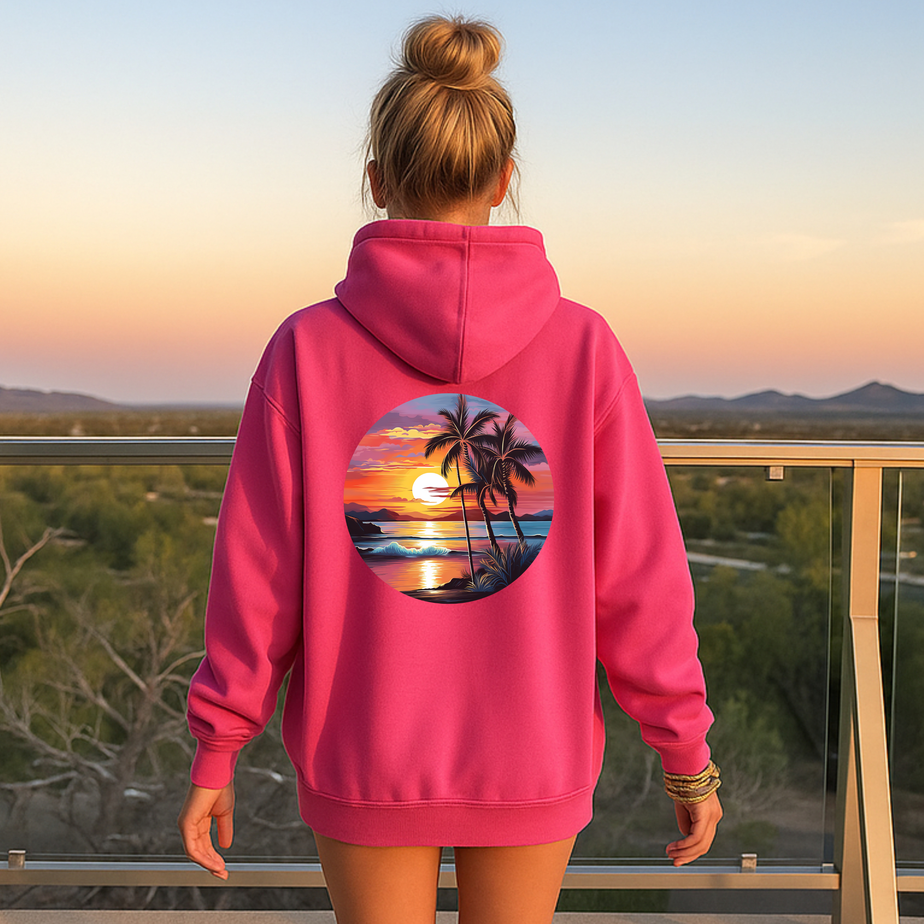 Person wearing a pink hoodie with a palm tree and sunset design, standing on a balcony with a scenic view.