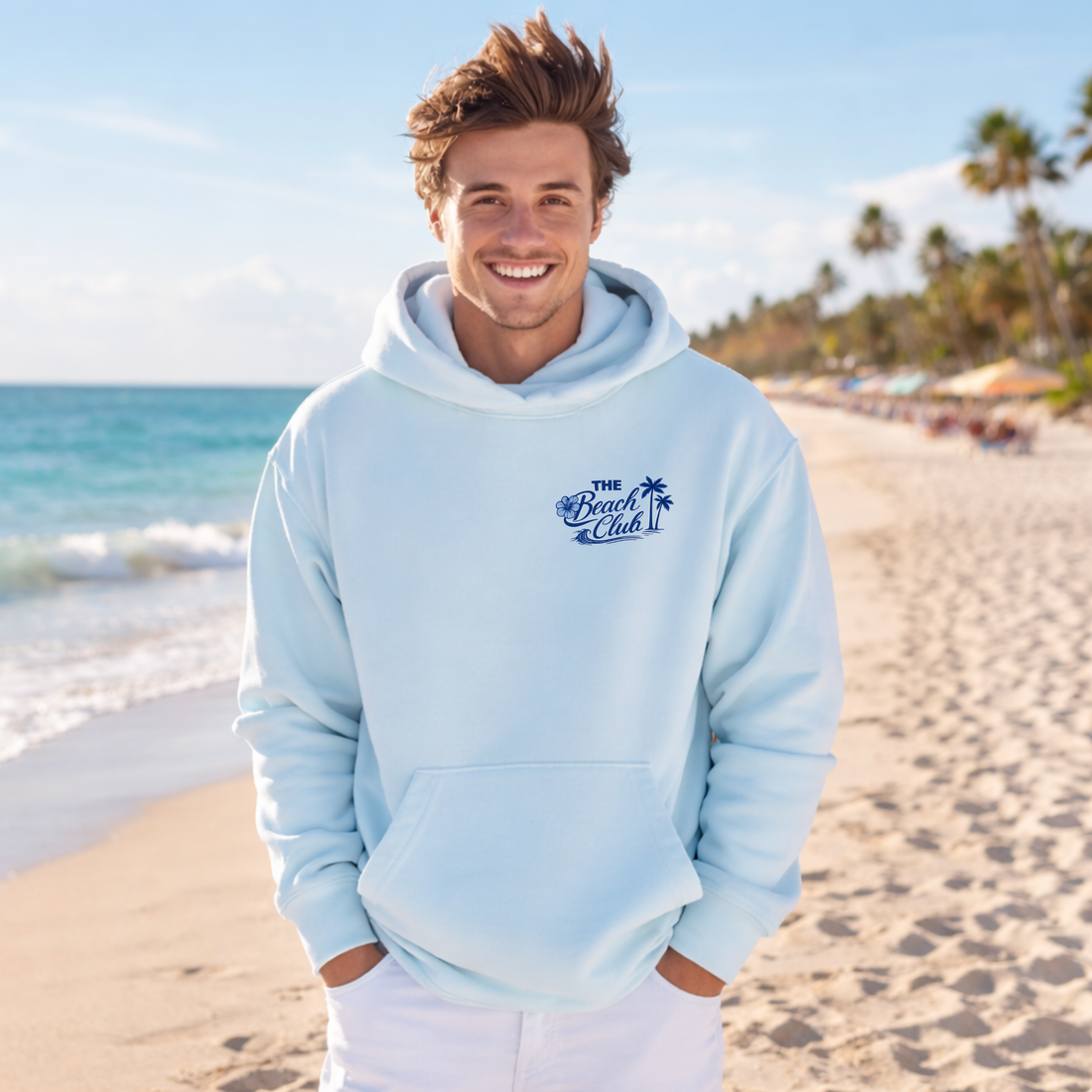 Person wearing a light blue hoodie with 'The Beach Club' design on a beach.